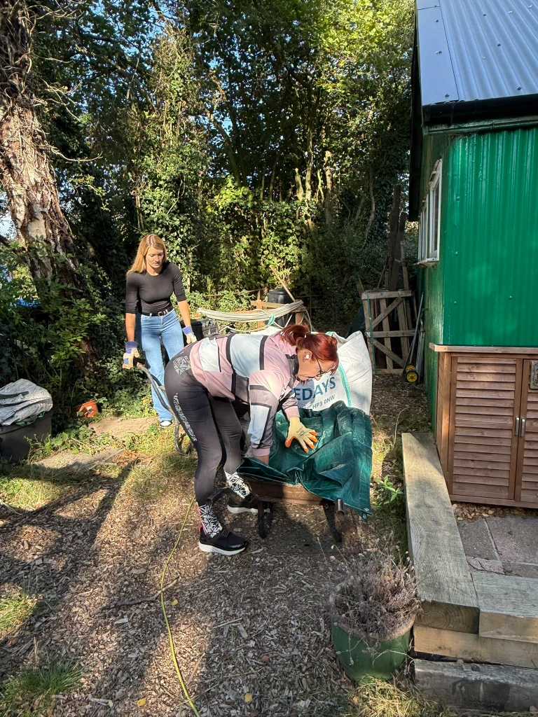 Volunteers loading garden waste onto a wheelbarrow beside the hut